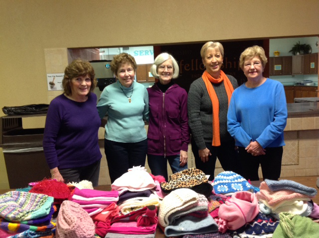 Five women standing behind a table filled with donated hats and mittens.